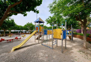 Legeplads med rutsjebane og klatrestativ på Camping La Masia feriepark, omgivet af træer og hegn.