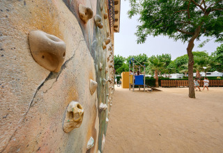 Klimmuur en speelplein aan Camping La Masia, vakantiepark in Catalonië, Spanje, met bomen en zand.