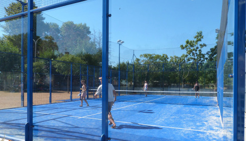 Personas jugando pádel en una pista azul al aire libre con paredes de vidrio en Camping La Masia, Cataluña.