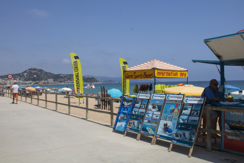 Paseo marítimo en Camping La Masia, Cataluña, con puesto de deportes acuáticos y sombrillas coloridas.