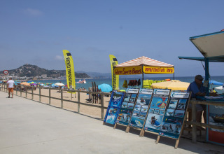 Boulevard bij Camping La Masia in Catalonië met kraam voor watersport en kleurrijke parasols op het strand.