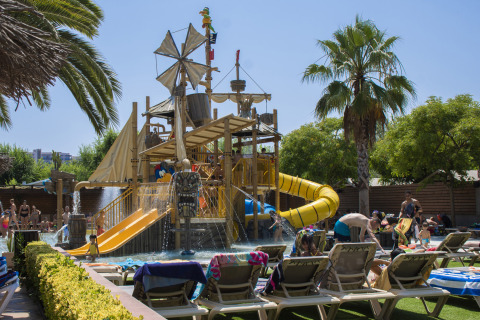 Niños jugando en un parque acuático con temática pirata, toboganes y palmeras en Camping La Masia, Cataluña, España.