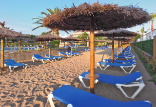 Area sabbiosa vuota con lettini blu e ombrelloni di paglia al Camping La Masia in Catalogna, Spagna.