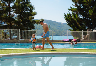 En far og hans lille barn går ved en udendørs swimmingpool i en feriepark med udsigt over bjerge og sø.