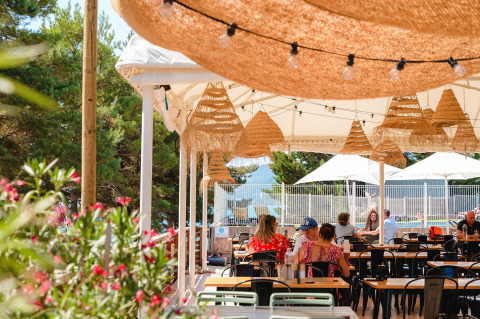 Udendørs café med stråtag og glødepærer, folk sidder ved borde, grønne træer og blomster i baggrunden.
