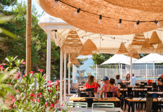 Udendørs café med stråtag og glødepærer, folk sidder ved borde, grønne træer og blomster i baggrunden.