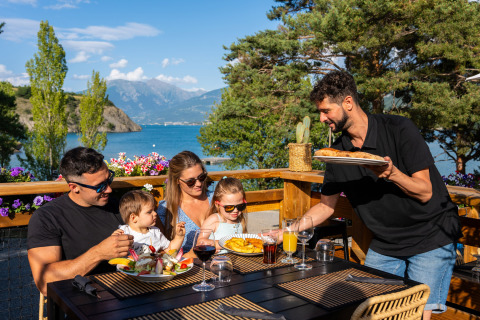 Familia disfruta de una comida al aire libre con vistas al lago y montañas en Camping La Presqu’île, Francia.
