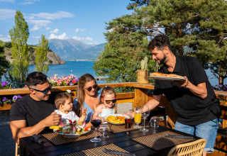 Familie genießt Mahlzeit auf Terrasse mit Blick auf See und Berge im Camping La Presqu’île, Frankreich.