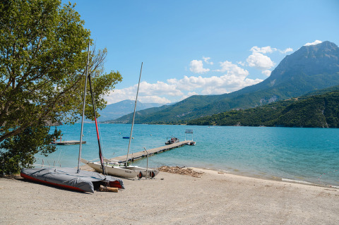 Scenic lake view with moored sailboats, a pier, and mountain backdrop at Camping La Presqu’île, France.
