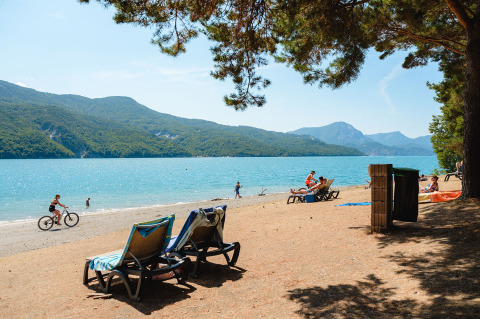 Escena de playa en Camping La Presqu’île en Provence-Alpes-Côte d’Azur, Francia, con tumbonas y montañas al fondo.