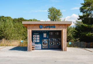 Buitenwasserette in houten gebouw bij Camping La Presqu’île, Provence-Alpes-Côte d’Azur, Frankrijk, met bomen rondom.