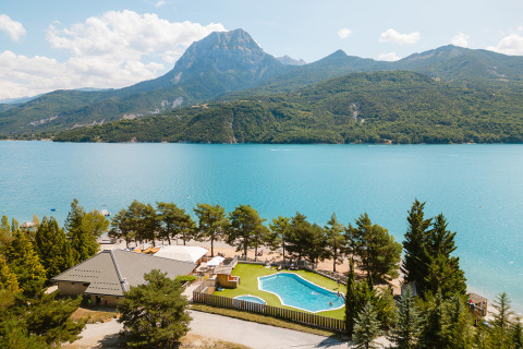 Parque de vacaciones con piscina junto a un lago turquesa y montañas al fondo en Provenza-Alpes-Costa Azul, Francia.