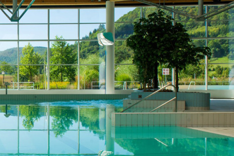 Indoor swimming pool with large windows overlooking green hills at Camping La Roche d’Ully, France.