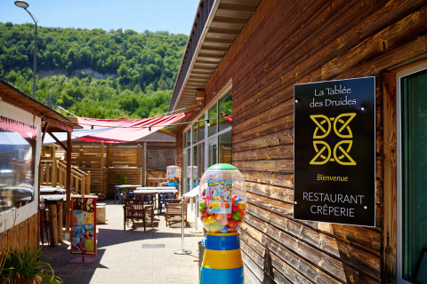 Outdoor dining area at Camping La Roche d’Ully with wooden buildings and forested hills in the background.