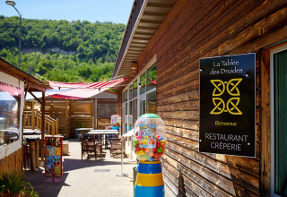 Udendørs spiseområde ved Camping La Roche d’Ully med træbygninger og skovklædte bakker i baggrunden.