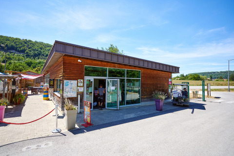 Entrance of Camping La Roche d’Ully, modern wooden building, outdoor seating, golf cart, and green hills.