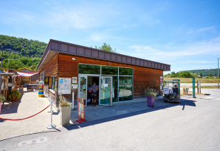 Entrance of Camping La Roche d’Ully, modern wooden building, outdoor seating, golf cart, and green hills.