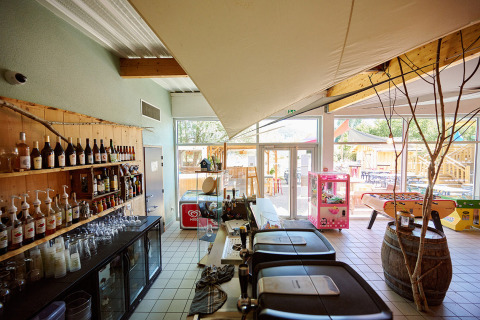 Zona de bar interior en Camping La Roche d’Ully con bebidas, juegos y vista a la terraza exterior.