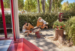 Two people are playing on a seesaw at a playground surrounded by trees in Camping Laguna Blu, Sardinia.