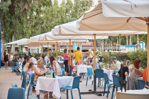 Area pranzo all'aperto al Camping Laguna Blu, Sardegna, Italia, con tavoli e ospiti sotto ombrelloni.