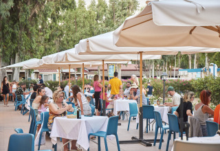 Area pranzo all'aperto al Camping Laguna Blu, Sardegna, Italia, con tavoli e ospiti sotto ombrelloni.