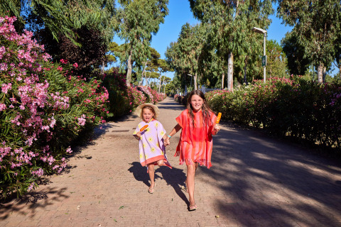 Twee kinderen wandelen vrolijk met ijsjes op een zonnig pad tussen bloeiende struiken in Sardinië.