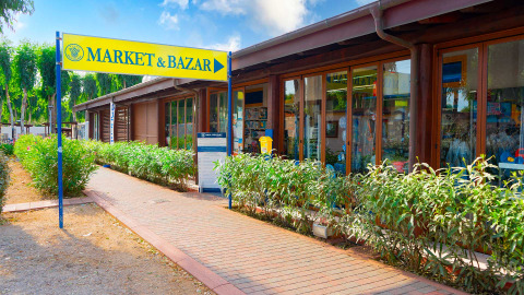 Ingresso al MARKET & BAZAR presso Camping Laguna Blu, Sardegna, con negozi e piante verdi sul vialetto.