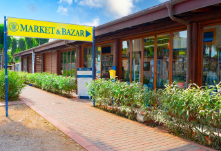 Ingresso al MARKET & BAZAR presso Camping Laguna Blu, Sardegna, con negozi e piante verdi sul vialetto.