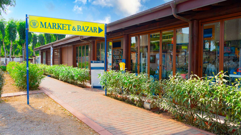 Eingang zum MARKET & BAZAR beim Camping Laguna Blu, Sardinien, mit Geschäften und Pflanzen draußen.