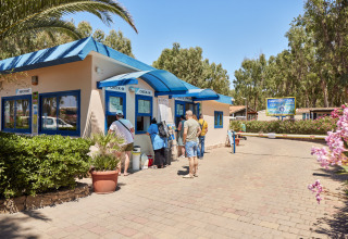 Ferieparkgæster venter ved Camping Laguna Blus check-in skranke i Sardinien, Italien, omgivet af grønt.