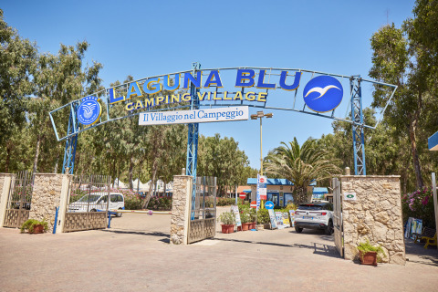 Cartel de entrada al camping Laguna Blu en Cerdeña, Italia, con coches y árboles al fondo.