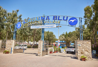 Cartel de entrada al camping Laguna Blu en Cerdeña, Italia, con coches y árboles al fondo.