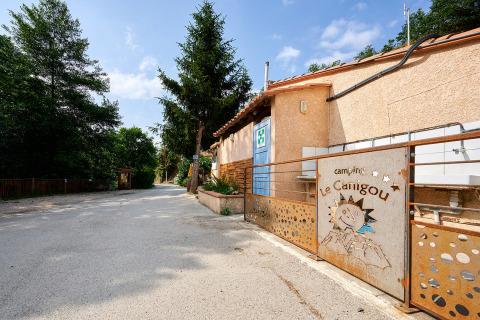 Entrance of Camping Le Canigou holiday park in Occitanie, France, featuring trees, a blue sky and a rustic gate.