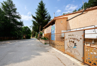 Indgang til Camping Le Canigou i Occitanie, Frankrig, med bygninger, træer og rustik port under blå himmel.