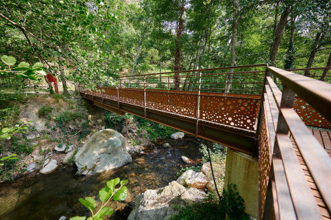 Puente peatonal metálico sobre arroyo en Camping Le Canigou, Occitania, Francia, rodeado de árboles.