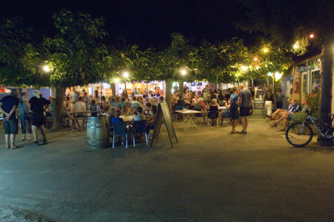 Outdoor evening scene with people enjoying food and drinks under trees at Camping Le Canigou in Occitanie, France.
