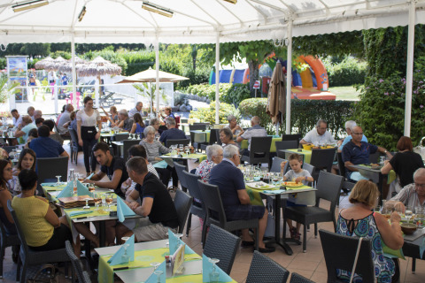 Terraza del restaurante en Camping Le Coin Tranquille con comensales y zona de juegos en el fondo.