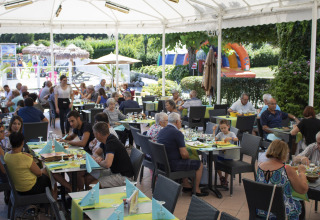 Outdoor dining area at Camping Le Coin Tranquille, guests eating and play area visible in the background.