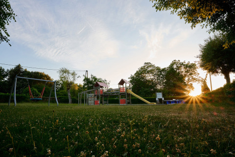 Parco giochi al Camping Le Coin Tranquille, Auvergne-Rhône-Alpes, Francia, con altalene, scivolo e tramonto sullo sfondo.