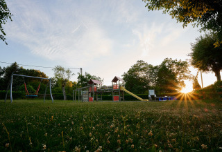 Parco giochi al Camping Le Coin Tranquille, Auvergne-Rhône-Alpes, Francia, con altalene, scivolo e tramonto sullo sfondo.