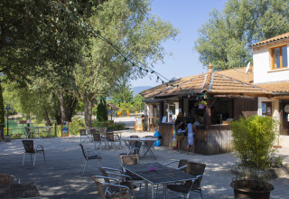 Buitenterras met tafels, stoelen en veel bomen op Camping Le Lac Bleu in Auvergne-Rhône-Alpes, Frankrijk.