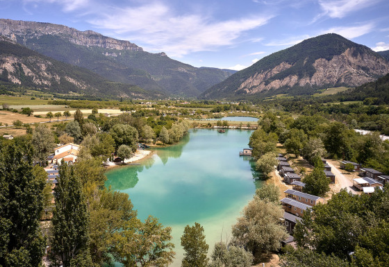 Veduta panoramica di Camping Le Lac Bleu in Alvernia-Rodano-Alpi, Francia, con lago, chalet e montagne.