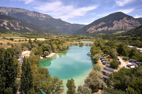 Veduta panoramica di Camping Le Lac Bleu in Alvernia-Rodano-Alpi, Francia, con lago, chalet e montagne.