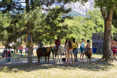 Niños y adultos con ponis bajo los árboles en Camping Le Lac Bleu, parque de vacaciones en Auvergne-Rhône-Alpes, Francia.