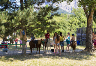 Niños y adultos con ponis bajo los árboles en Camping Le Lac Bleu, parque de vacaciones en Auvergne-Rhône-Alpes, Francia.