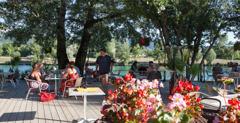 Outdoor café terrace by the lake at Camping Le Lac Bleu in Auvergne-Rhône-Alpes, France, with guests and flowers.