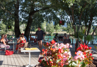 Outdoor café terrace by the lake at Camping Le Lac Bleu in Auvergne-Rhône-Alpes, France, with guests and flowers.