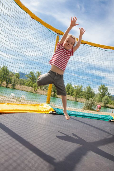 Niño saltando en una cama elástica junto al lago en Camping Le Lac Bleu, parque de vacaciones en Auvernia-Ródano-Alpes, Francia.