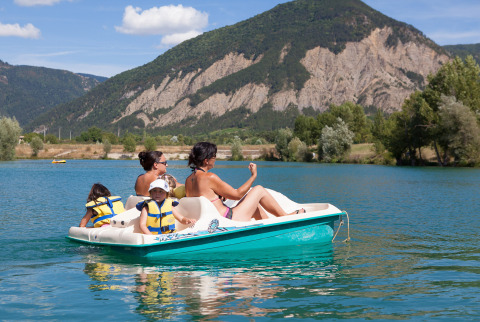 Family enjoying a pedal boat ride on a lake at Camping Le Lac Bleu with mountains in Auvergne-Rhône-Alpes.