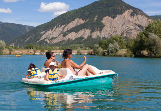 Family enjoying a pedal boat ride on a lake at Camping Le Lac Bleu with mountains in Auvergne-Rhône-Alpes.